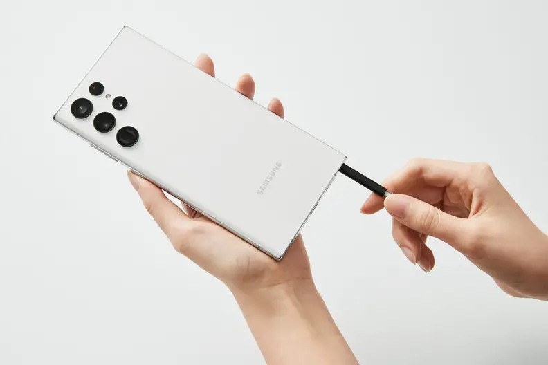 A white Samsung S22 Ultra being held in a white person's hand in front of a white background. Their other hand is removing the stylus.