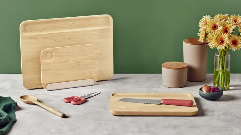 Serrated knife sitting on top of wooden cutting block sitting next to bowl of dates, vase of sunflowers, a pair of scissors, a wooden spoon, two wooden cutting blocks and two storage containers.