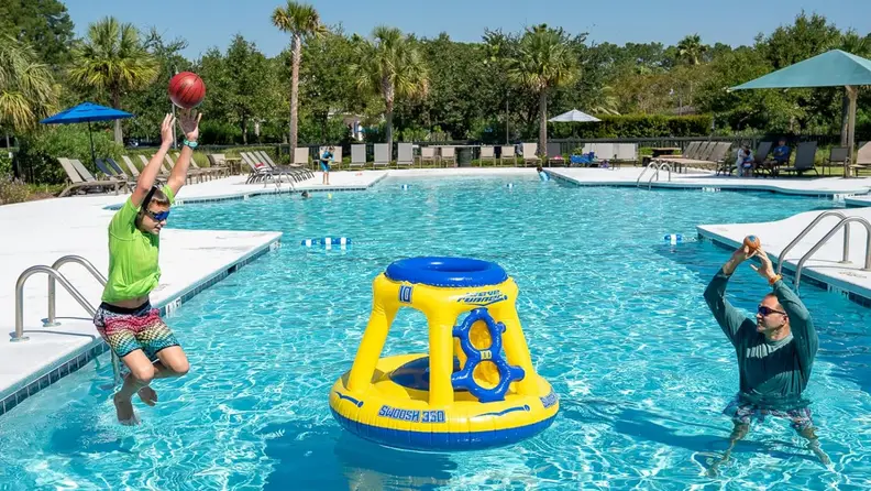 A youth jumps into a pool while shooting a ball at the Wave Runner Swoosh 360 pool basketball hoop as a person in the pool throws a ball toward the hoop.
