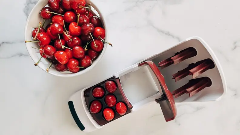 A bowl of cherries sits beside the OXO multi-cherry pitter on a marble surface.