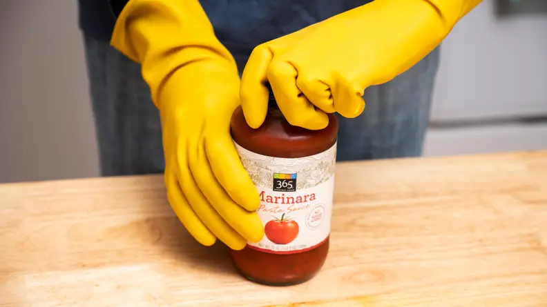 A pair of hands wearing yellow rubber gloves opening a glass jar of 365 marinara sauce.