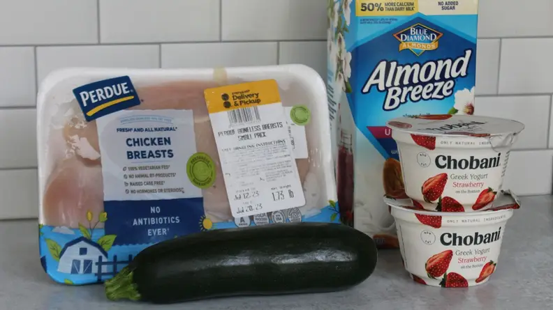 Pack of raw chicken breasts, uncut cucumber, two cups of Chobani strawberry yogurt and a box of almond milk sits together on kitchen counter in front of tile wall.
