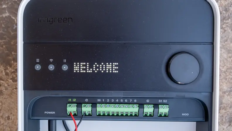 Actual Irrigreen Smart Sprinkler unit shown in black with a welcome screen and various wire ports.