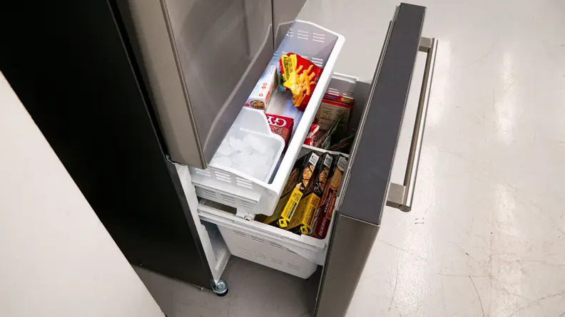 A side view of the fridge's open freezer drawer. We can see the main bin stored with frozen food. The shelf above holds the reservoir for the ice maker and has some more food stored beside it.