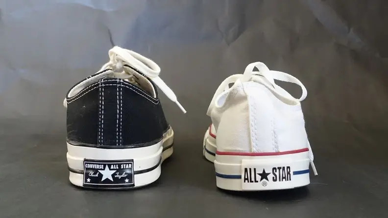 On the left, a Chuck 70 low top sneaker in black. On the right, a white Chuck Taylor All Star low top sneaker. Both sneakers feature different logos on their heels.