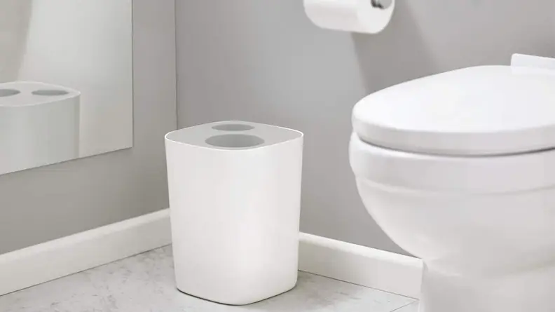 A all-gray bathroom contains a white toilet, a roll of toilet paper, and a trash can with an opening for trash and an opening for recycling.