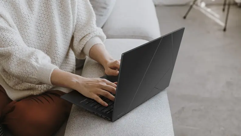 A person in a white sweater types on an Asus Zenbook 14 OLED UM3406HA while sitting on a white sofa.