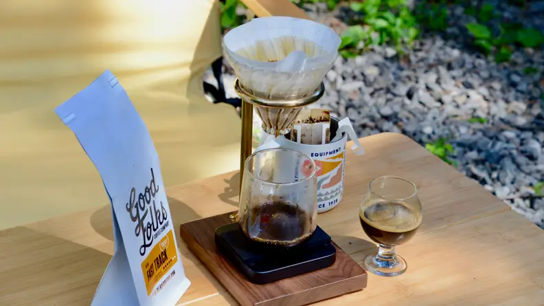 A pour-over coffee maker sits on a wooden table outside, accompanied by a bag of coffee beans and a cup filled with freshly made espresso.