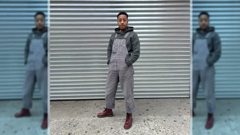 A person wearing overalls, red boots, and a gray hoodie, standing in front of a metal background.