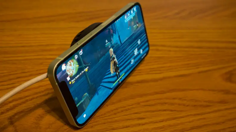 An angled shot of an iPhone 12 Mini, with the fan attached, being propped up by the Phone Cooler, with Genshin Impact showing on screen.
