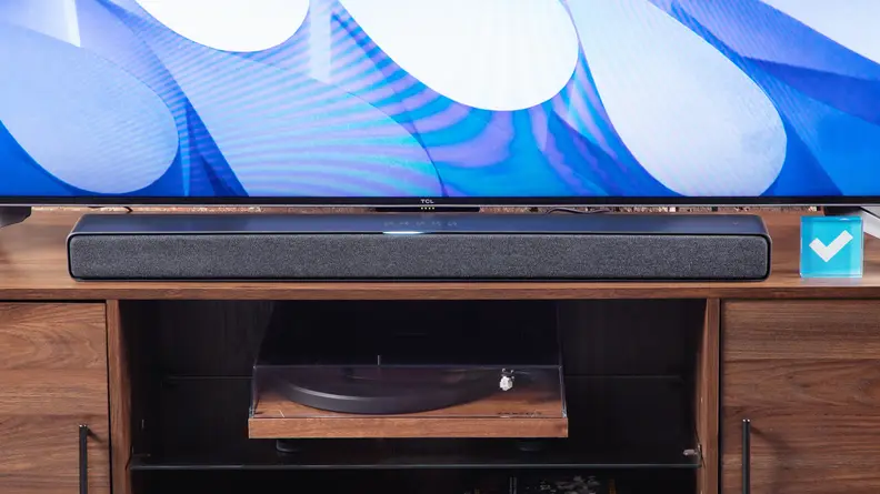 TCL Q Class 3.1 Soundbar sitting on top of wooden credenza in front of flat screen television and acrylic paper weight with blue checkmark in the middle.