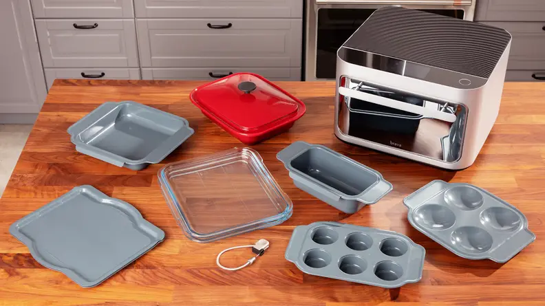 The Brava Glass Oven on a wooden countertop next to assorted cooking trays and dishes.