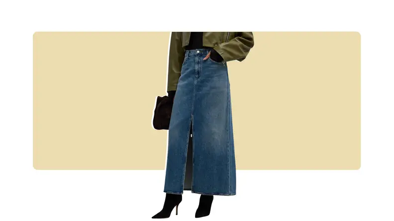 A model seen from the waist down wearing an ankle-length blue denim skirt with a front slit, paired with black boots, an olive jacket, and a bag.