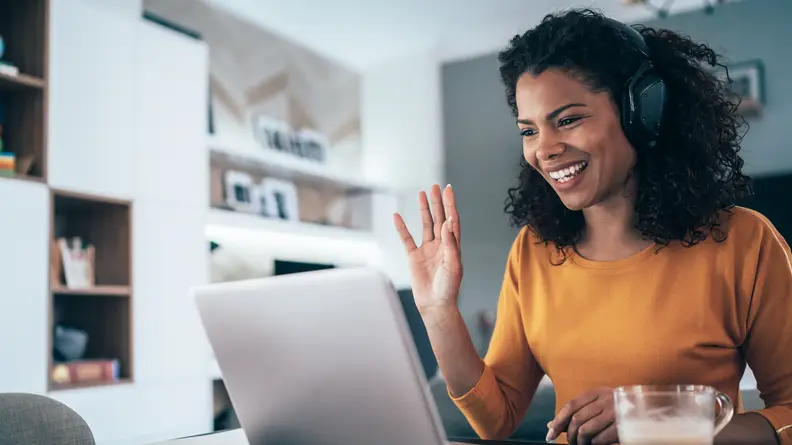 Woman in yellow shirt waves to computer in home office