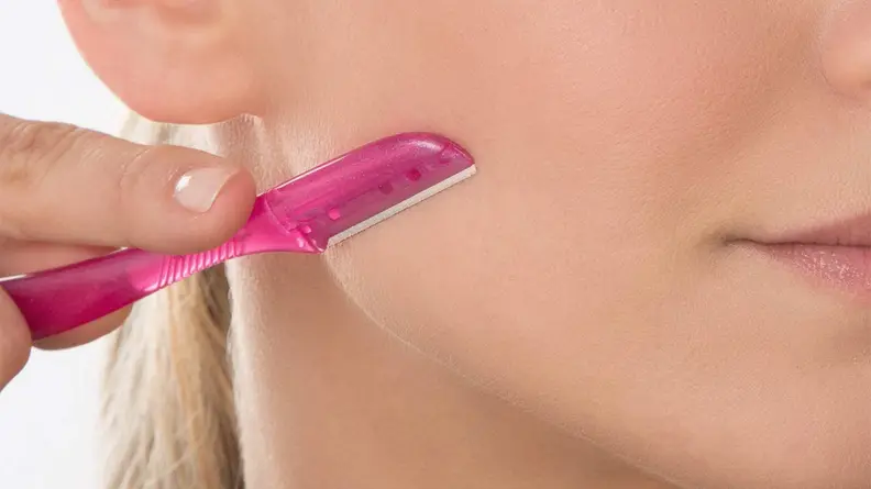 A closeup of a woman holding a dermaplane tool on her jawline pointing downward.