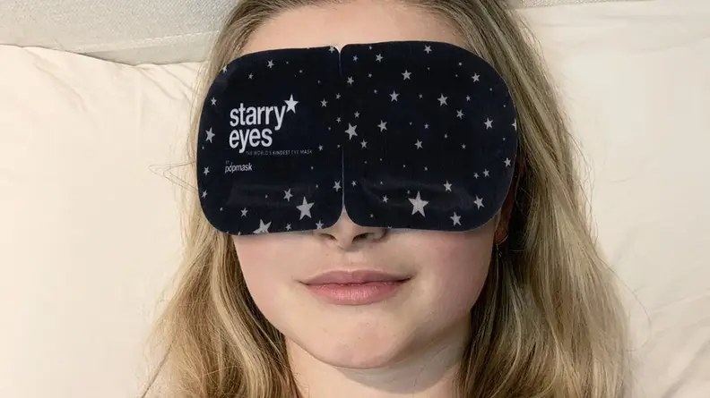A person with long blonde hair wears a large black eyemask.