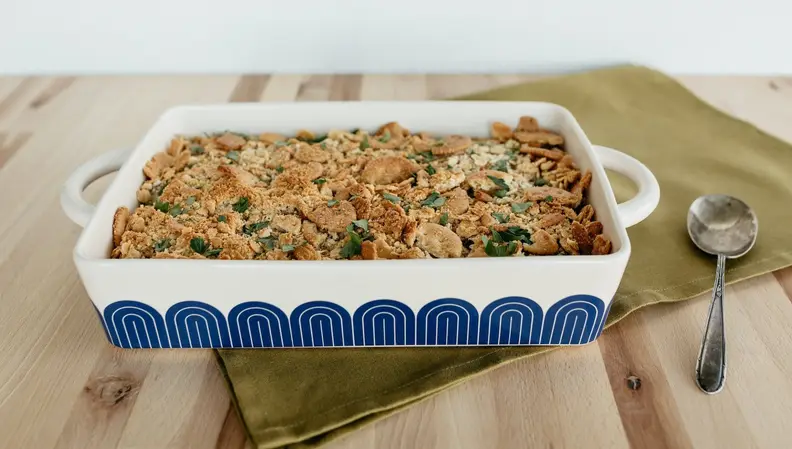 A blue and white Great Jones Hot Dish that's filled with a casserole sits on a wood table.