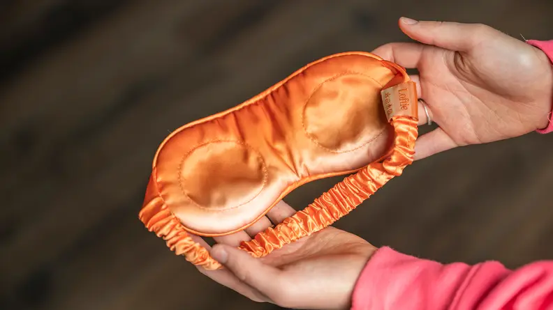 Person holding orange satin sleep mask in hands.