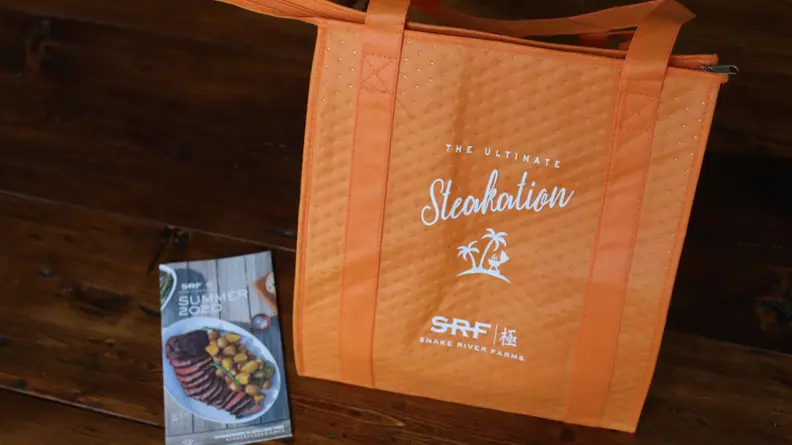 The Snake River Farms orange tote bag is displayed in the center of the image, accompanied by a brochure from the same business.