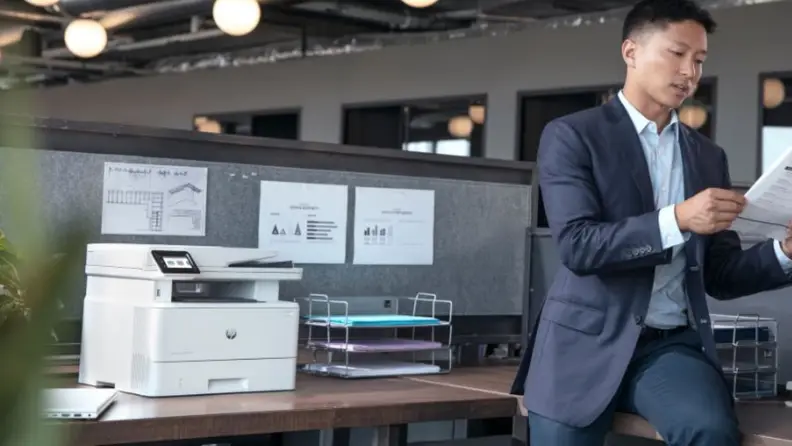 A man in a suit reviews documents while leaning against a desk next to an HP LaserJet Pro MFP 4101fdw.