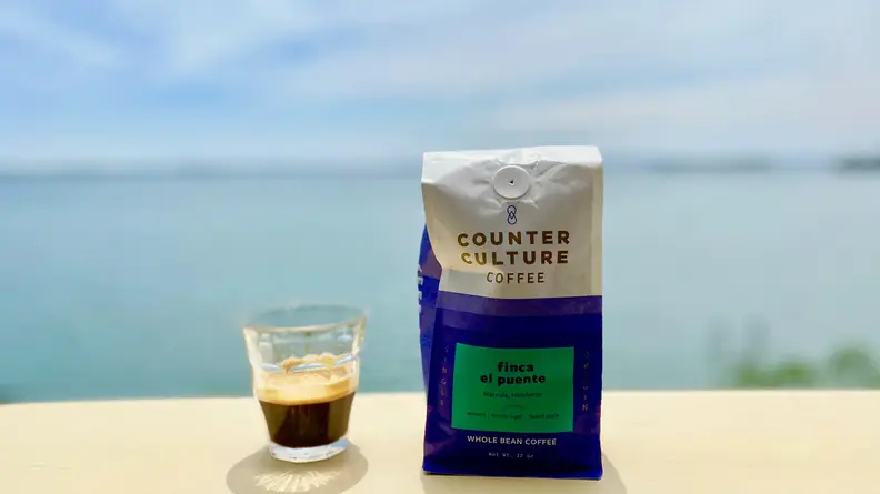 A glass espresso cup filled with a shot of espresso sits next to a bag of coffee beans on a table outside.