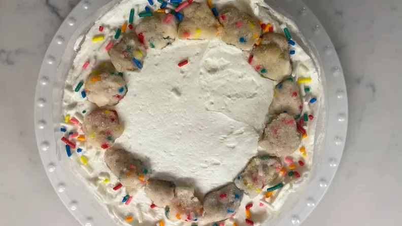 A top-down photo of a Milk Bar birthday cake with white icing and sprinkled cookie dough chunk garnish.