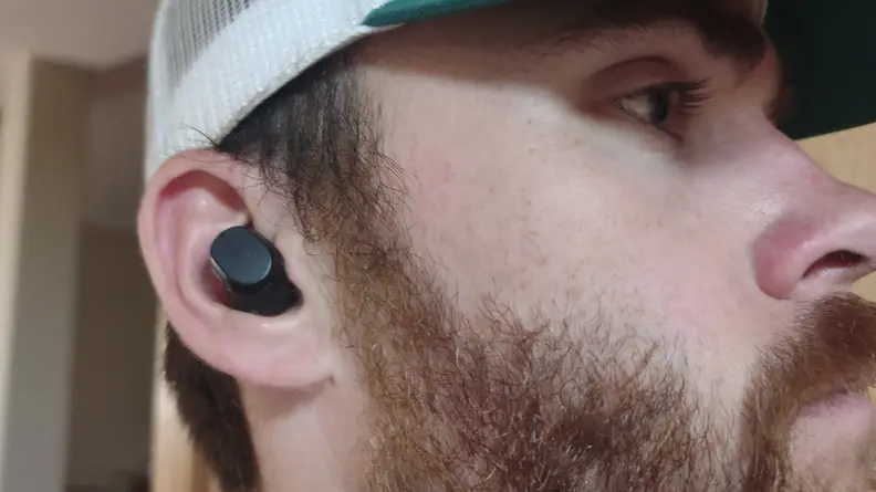 The all-black Skullcandy Mod earbuds in the ear of a person wearing a hat with a red beard.