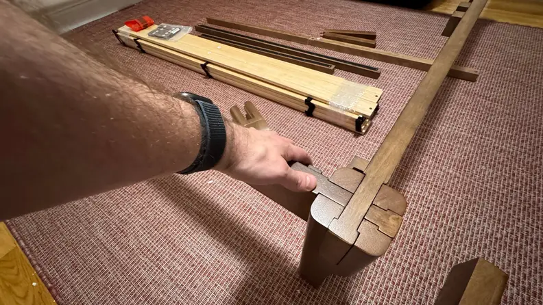 Disassembled parts of the FlexiSpot Kana bed frame, with a left arm touching the frame on top of a maroon and white rug.