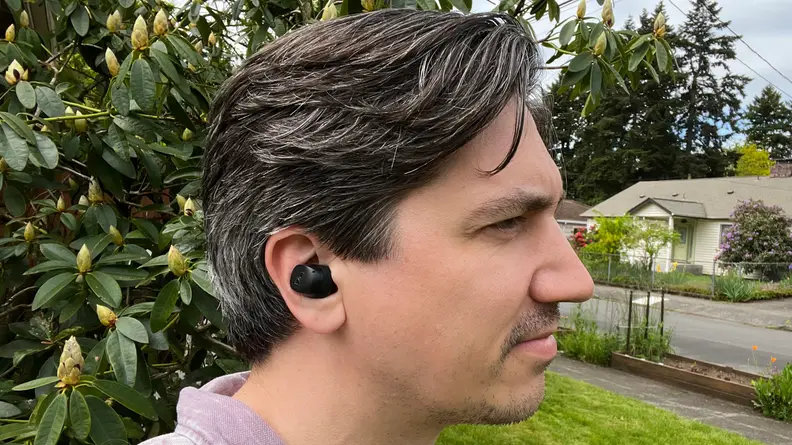 A black model of Sennheiser's Momentum TW 3 earbuds sit in a man's ear in front of green bushes and a green lawn.