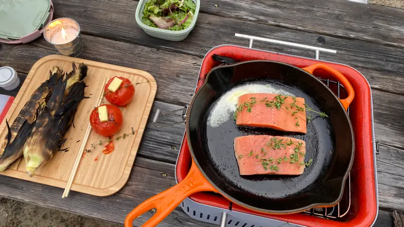 An orange cast iron skillet is placed on top of the Everdure Cube charcoal grill. In the skillet, there are two pieces of salmon. Next to the grill, there's a bamboo tray of cooked corn and tomatoes.