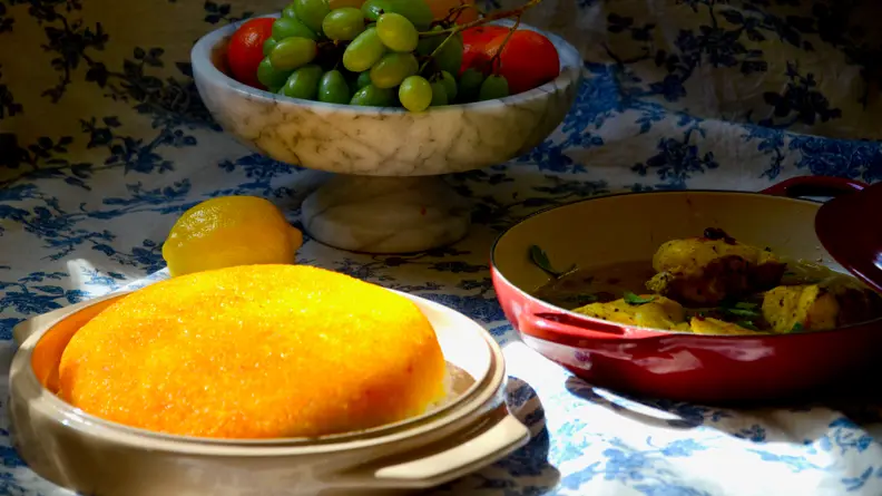 A plate of golden tahdig in the noosh-e-joon platter is in the center of the image. In the shadow, there's a plate of fruits and a pot of saffron chicken.
