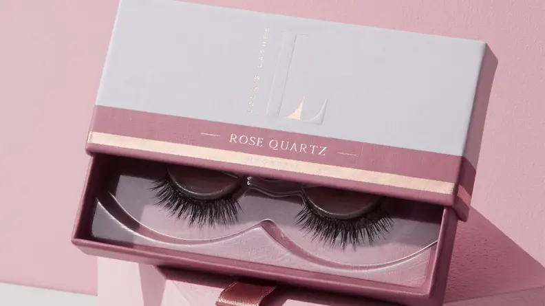 The pair of lashes from the Lola’s Lashes Rose Quartz Magnetic Eyelashes and Eyeliner Kit sitting in its packaging on a pink background.