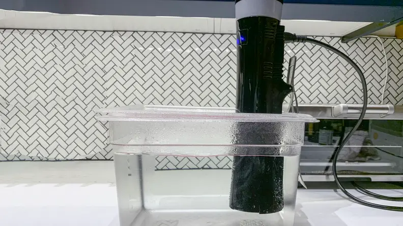 A sous vide immersion circulator attached to the side of a plastic tub that's been partially filled with water.