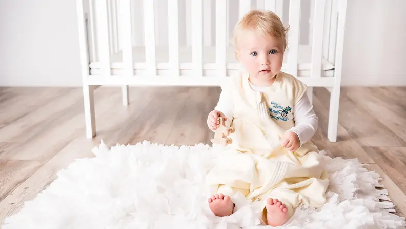 A baby wearing a cream onesie sits in front of a crib.