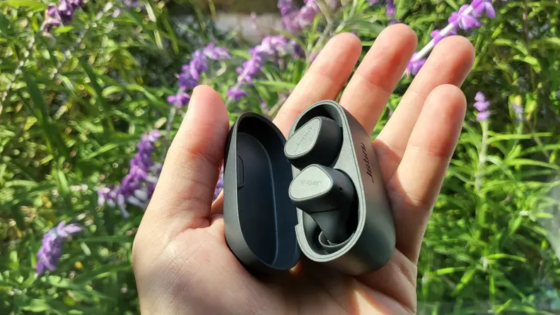 Jabra's all-black, matte earbuds sit in their open case in an open hand above green and purple flowers blown in a light breeze.