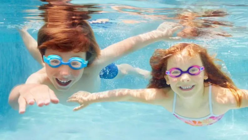 Two kids swim under water smiling in their swimming goggles