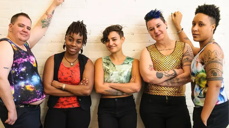 A group of people wearing binders of different patterns and designs, all wearing black pants, standing up against a wall.