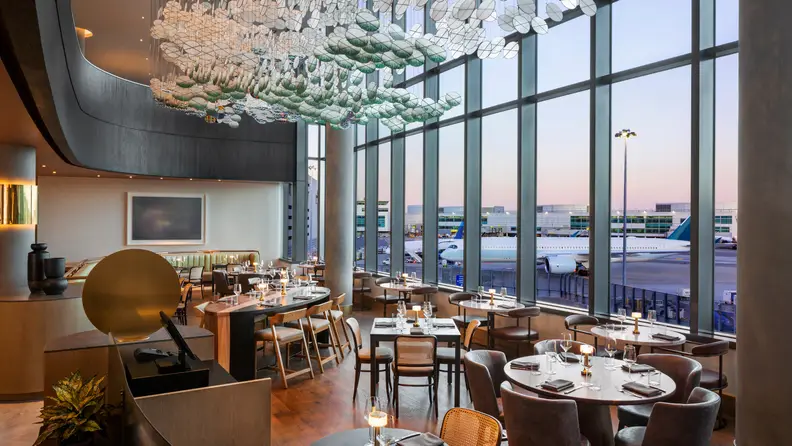 Upscale airport restaurant with floor-to-ceiling windows overlooking planes on the runway, featuring modern décor, candlelit tables, and a sculptural ceiling installation.