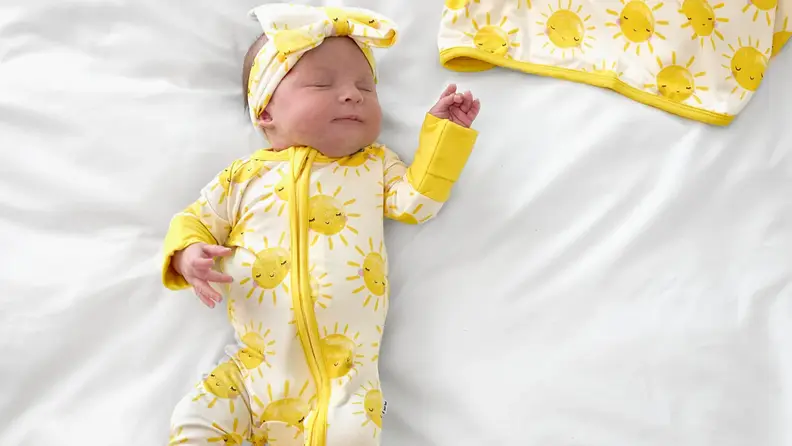 A sleeping baby wearing a yellow sun-print onesie and matching headband lies on a white bed.