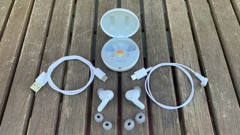 The LG T90Q earbuds with two extra sets of ear tips, two USB-C cables, and their open case all sitting on a wooden table outdoors.