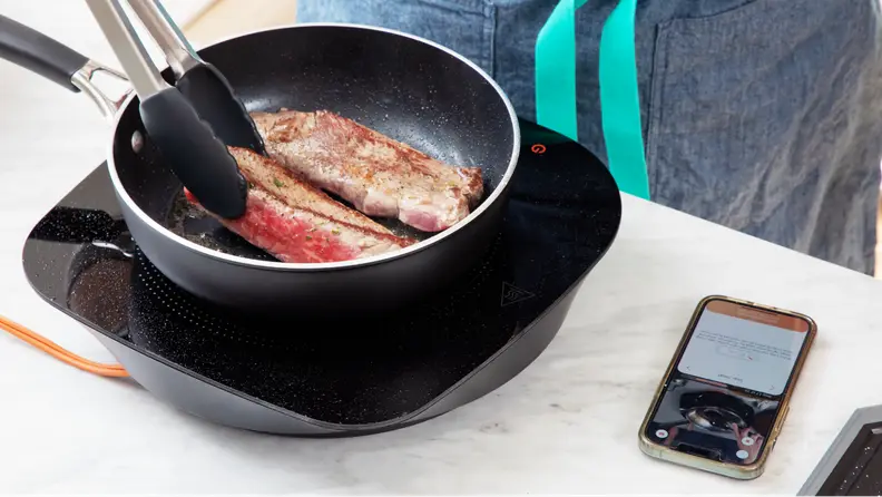Two pieces of steak being cooked atop cookware with a smartphone displaying a recipe.