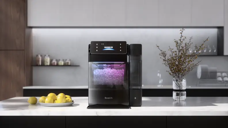 A countertop ice maker sits in a modern kitchen, with a clear window showing freshly made ice illuminated inside the machine.
