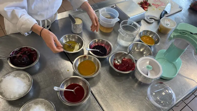 A variety of shaved ice toppings including condensed milk and flavored simple syrups, arranged on a stainless steel kitchen table.