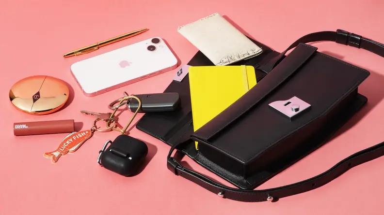 A black leather Behno handbag lays on a pink surface, the contents pouring out across the table, including a phone, pen, keys, makeup, and a journal.