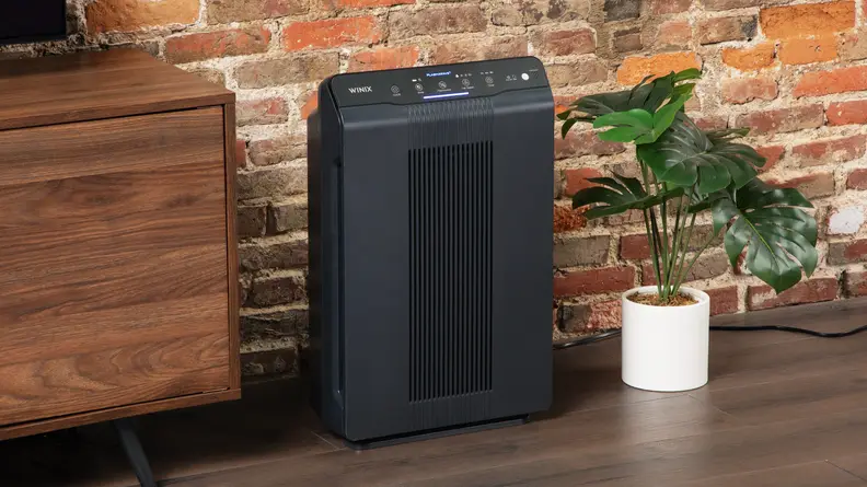 Black Winix 5500-2 air purifier on top of wooden floorboard in front of brick wall indoors.