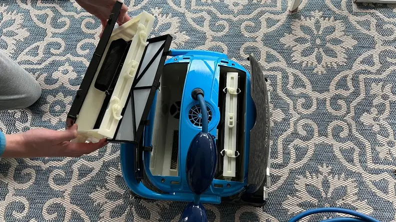 The Dolphin Nautilus CC Plus Wi-Fi  on an outdoor rug with a person holding one filter basket open and the other filter basket is exposed.