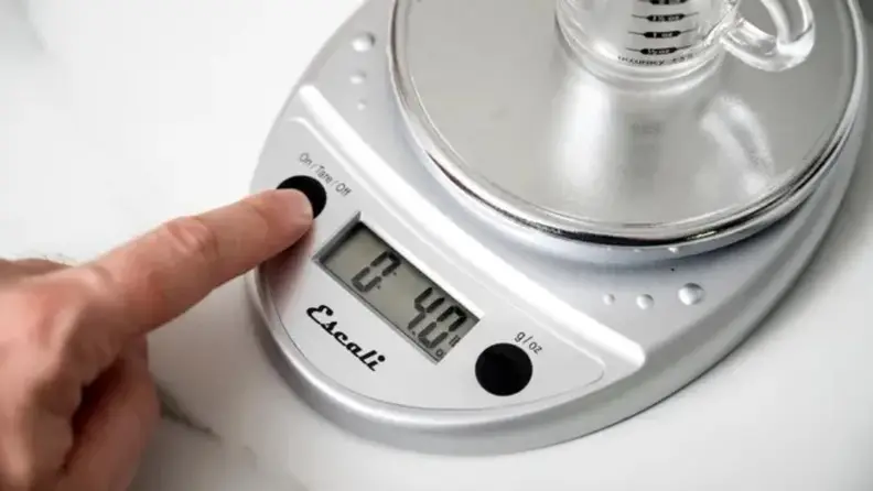 A hand presses a button on the Escali digital kitchen scale.
