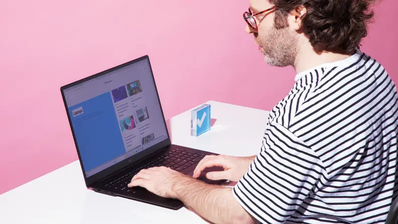 A man sitting in a chair, typing on the LG gram Pro 17 laptop on a white desktop with a pink background.