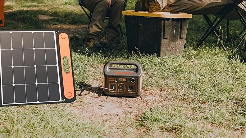 A power station sits on the ground in the woods, getting recharged by a nearby solar panel. In the background you can see two campers relaxing.