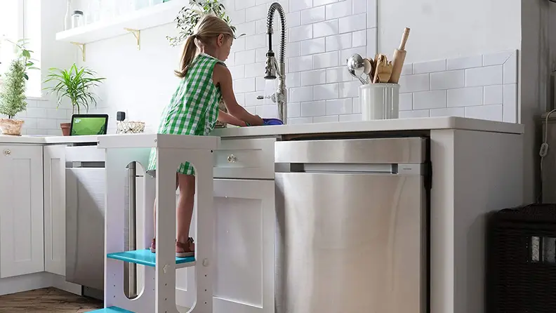 A child with a ponytail standing on the Little Partners Explore n Store learning tower to reach a kitchen sink